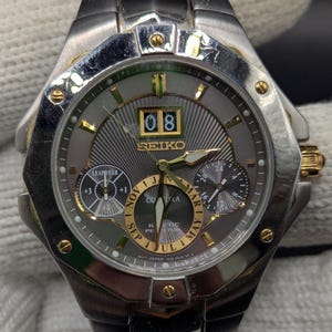 May include: A silver and gold-toned Seiko watch with a gray face. The watch features a date display, multiple sub-dials, and gold accents on the bezel and band. The watch is displayed on a white surface.