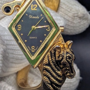 May include: A gold-toned quartz watch with a black face and a diamond-shaped bezel. The watch features the brand name "Xanadu" and a zebra head design on the band. The watch band is a cuff style.
