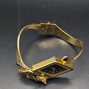 May include: A gold-toned bracelet watch with a black watch face. The watch face is square with gold-colored hour markers. The bracelet has a decorative leaf design and a clasp closure.