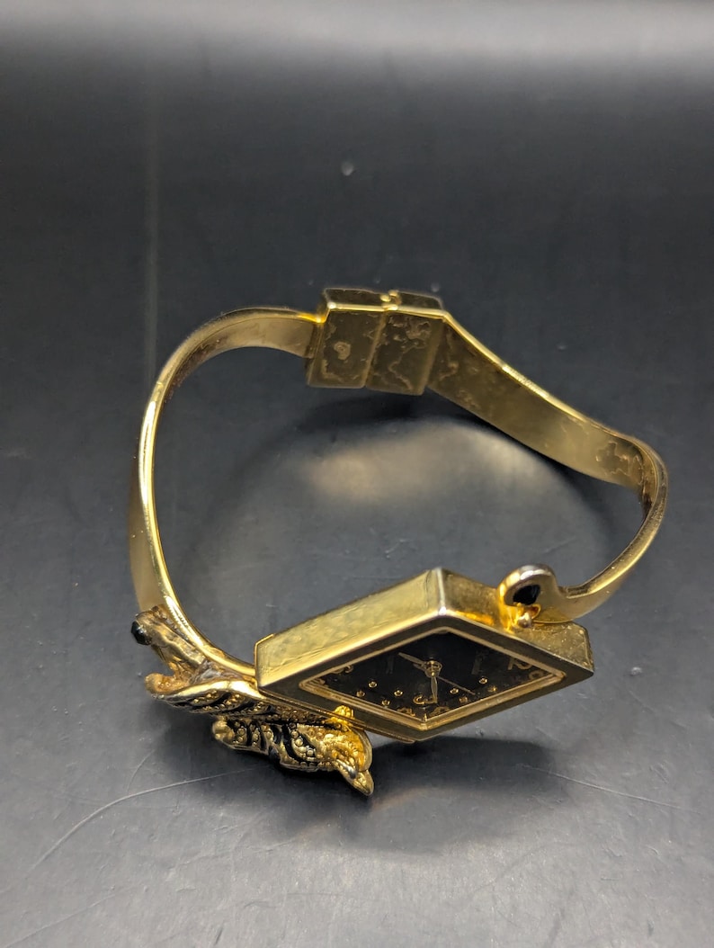 May include: A gold-toned bracelet watch with a black watch face. The watch face is square with gold-colored hour markers. The bracelet has a decorative leaf design and a clasp closure.