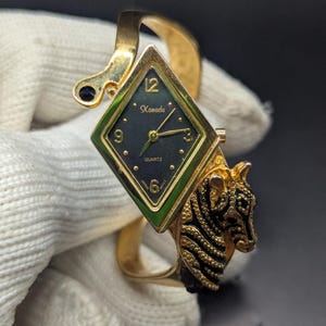 May include: Gold-toned quartz watch with a diamond-shaped face and a tiger head design. The watch face is dark green with gold hands and the word "Kanadu". The watch band is a gold cuff style.