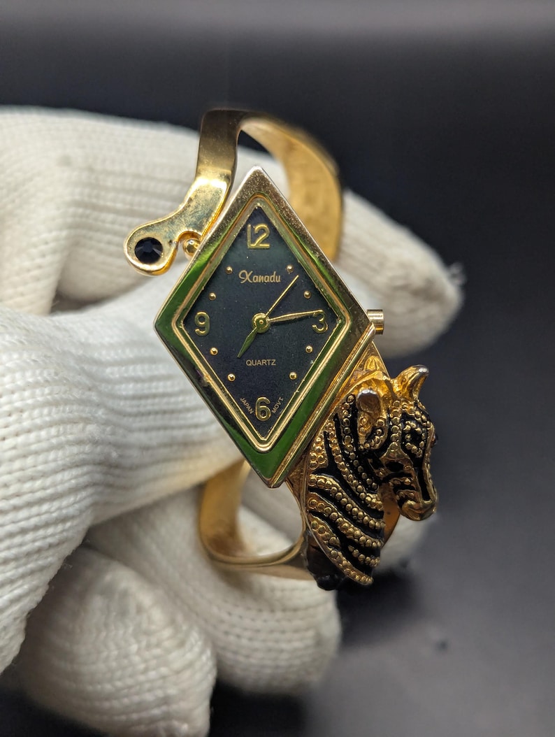 May include: Gold-toned quartz watch with a diamond-shaped face and a tiger head design. The watch face is dark green with gold hands and the word "Kanadu". The watch band is a gold cuff style.