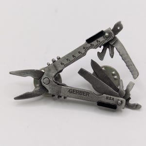 Pode incluir: Um alfinete multi-ferramenta prateado com a marca "GERBER" e "U.S.A." impressas. A ferramenta inclui alicates, uma régua, uma faca e outros implementos. O alfinete foi projetado para se parecer com uma versão em miniatura de uma multi-ferramenta de tamanho normal.