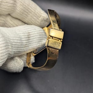 May include: A gold-colored bracelet with a rectangular clasp and a curved band. The bracelet is held by a gloved hand against a dark background. The clasp has a hinged design.