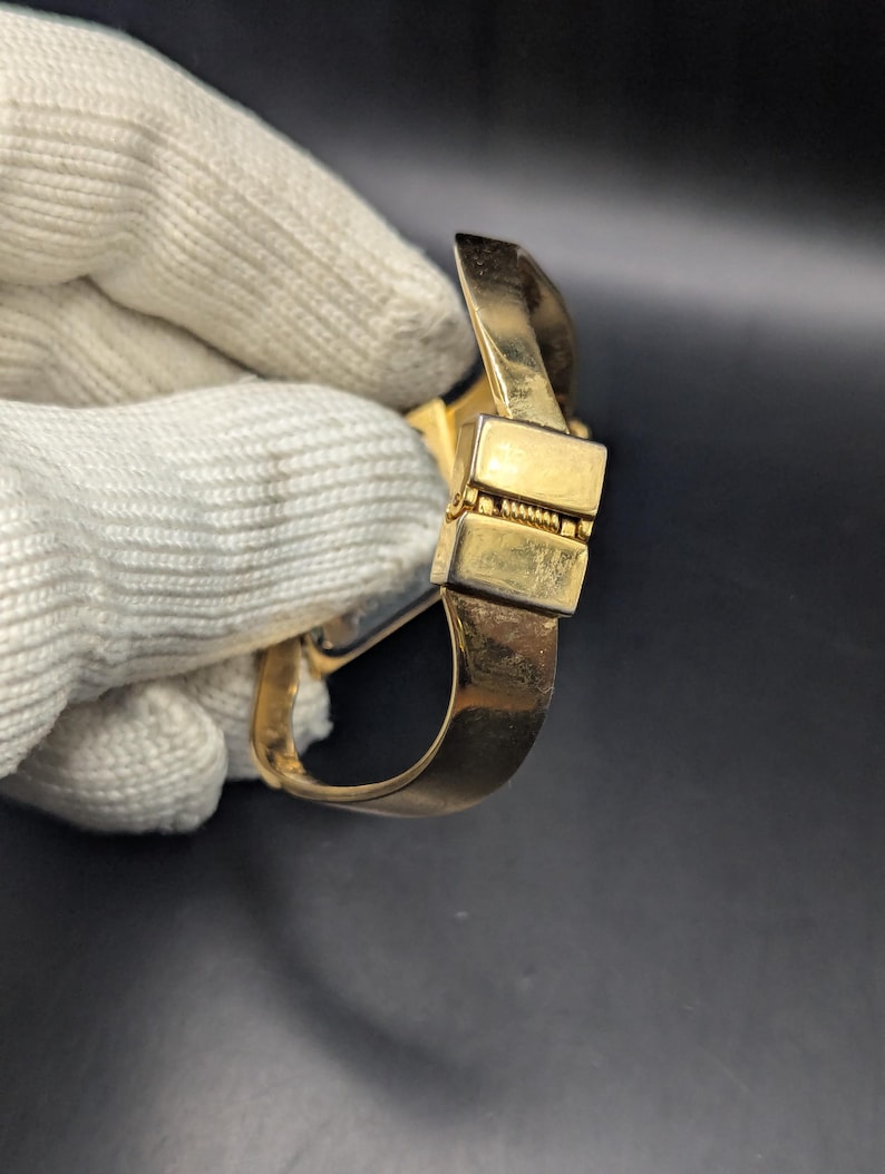 May include: A gold-colored bracelet with a rectangular clasp and a curved band. The bracelet is held by a gloved hand against a dark background. The clasp has a hinged design.