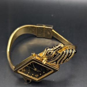 May include: A gold-toned wristwatch with a black face and a unique design. The watch features a diamond-shaped face with gold hands and hour markers. The band is gold, and the clasp is visible. The watch has a decorative black and gold animal head on the side.