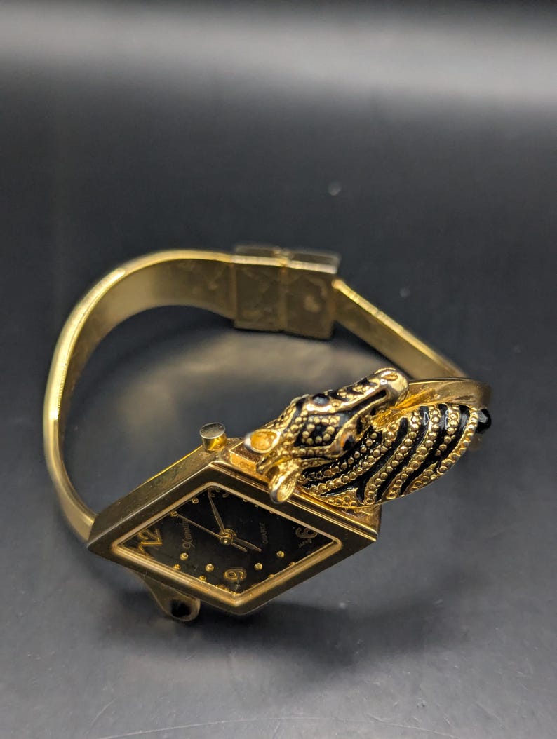 May include: A gold-toned wristwatch with a black face and a unique design. The watch features a diamond-shaped face with gold hands and hour markers. The band is gold, and the clasp is visible. The watch has a decorative black and gold animal head on the side.
