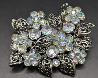 Peut inclure: Broche florale argentée ornée de strass clairs et irisés. La broche présente un motif ajouré détaillé de feuilles et de fleurs, avec des strass regroupés au centre de chaque fleur. Les strass ont un éclat arc-en-ciel.