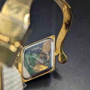 May include: A gold-toned Xanadu wristwatch with a rectangular face. The watch face has the words "Mineral Crystal" and "Japan Movement" printed on it. The watch band is a cream-colored fabric.