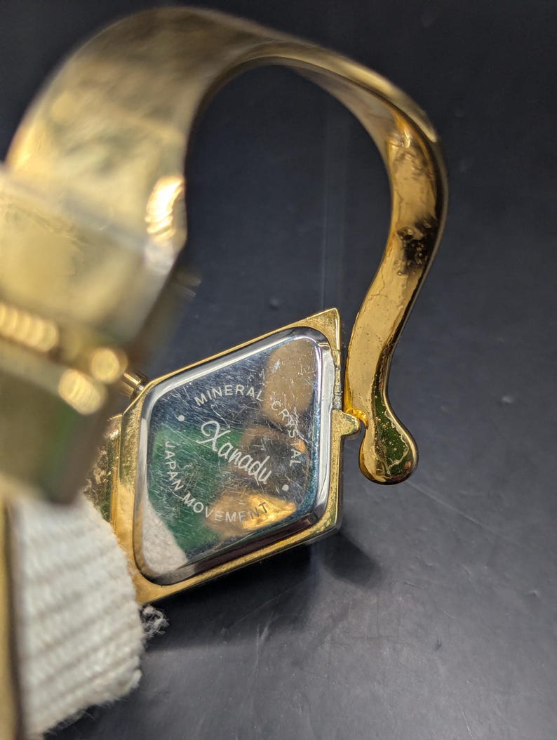 May include: A gold-toned Xanadu wristwatch with a rectangular face. The watch face has the words "Mineral Crystal" and "Japan Movement" printed on it. The watch band is a cream-colored fabric.