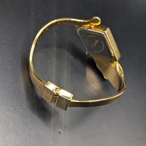 May include: Gold-toned wristwatch with a rectangular black watch face. The watch features a hinged bracelet-style band and a clasp closure. The watch face has the word "Kanada" printed on it.