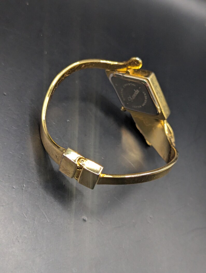 May include: Gold-toned wristwatch with a rectangular black watch face. The watch features a hinged bracelet-style band and a clasp closure. The watch face has the word "Kanada" printed on it.