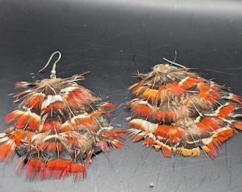 May include: A pair of feather earrings. Each earring features a cluster of layered feathers in shades of orange, red, brown, and white. The earrings have silver-colored hooks for pierced ears. The feathers are arranged in a fan-like shape.
