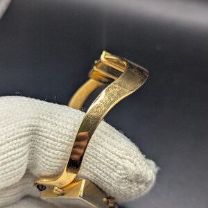May include: Close-up of a gold-colored bracelet with a rectangular clasp and a curved band. The bracelet is held by a white glove. The clasp has a small, dark stone.