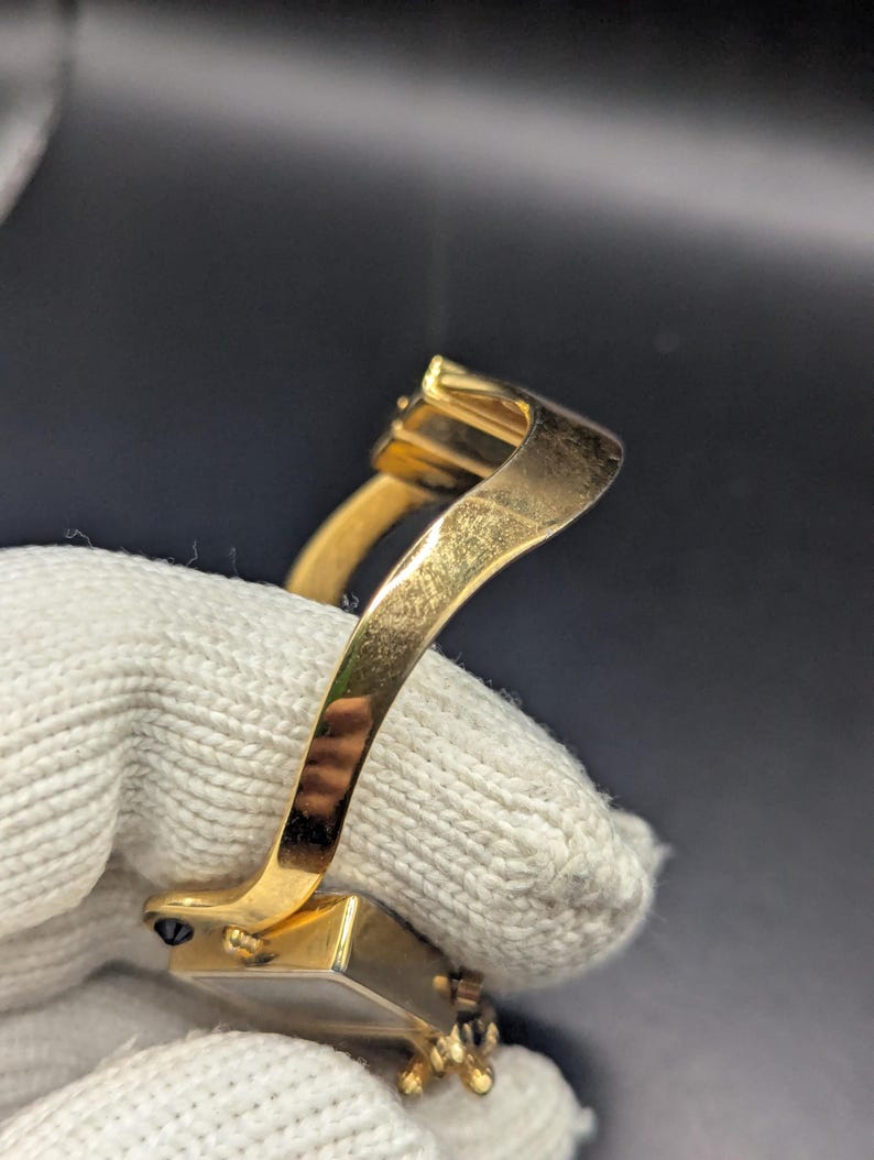 May include: Close-up of a gold-colored bracelet with a rectangular clasp and a curved band. The bracelet is held by a white glove. The clasp has a small, dark stone.