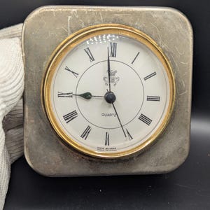 May include: Square, silver-toned quartz clock with a gold-colored bezel and a white face. The clock features black Roman numerals, black hands, and the word "QUARTZ" at the top. The clock reads 2:45. The words "MADE IN CHINA" are at the bottom.