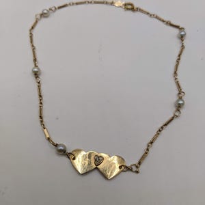 May include: A gold-toned necklace featuring three heart-shaped pendants with a small diamond accent. The necklace has a delicate chain design with small pearls interspersed throughout. The clasp is visible at the end of the chain.