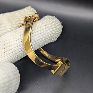 May include: A gold-colored bracelet with a rectangular clasp. The bracelet has a curved design and features small, dark-colored embellishments near the top. The bracelet is shown against a white fabric background.