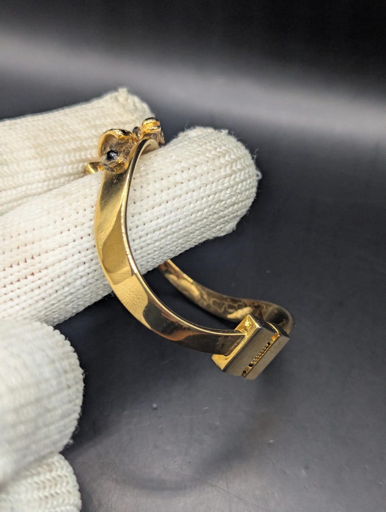 May include: A gold-colored bracelet with a rectangular clasp. The bracelet has a curved design and features small, dark-colored embellishments near the top. The bracelet is shown against a white fabric background.