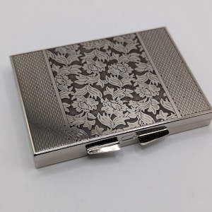 Puede incluir: Un estuche compacto plateado rectangular con un diseño floral en el centro y un patrón texturizado en los laterales. El estuche compacto tiene dos cierres plateados y un acabado brillante. El diseño es de estilo vintage.