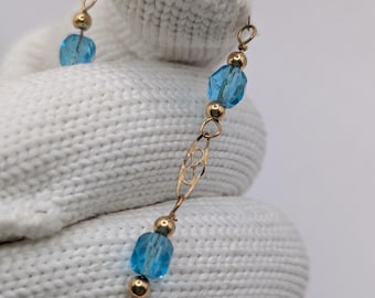 May include: A delicate gold necklace featuring light blue, faceted beads and small gold accents. The necklace is held against a white background, showcasing its intricate design and craftsmanship. The chain has decorative openwork links.