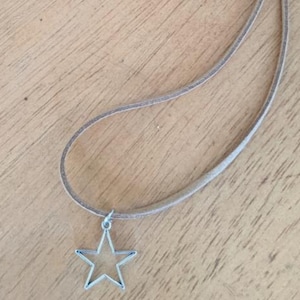 May include: A silver star pendant necklace with a light brown cord. The star is a simple, open design. The necklace is displayed on a wooden surface.