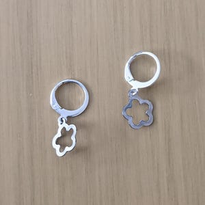 May include: A pair of silver-colored hoop earrings. Each earring features a small, openwork flower charm dangling from a hinged hoop. The earrings are displayed on a light brown wooden surface.