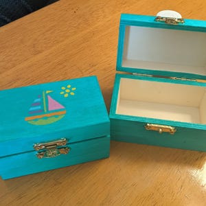 May include: Two small, turquoise wooden boxes with gold-colored clasps. One box is open, revealing a white interior. The closed box features a painted sailboat design with a pink sail and a yellow flower. The boxes are on a wooden surface.