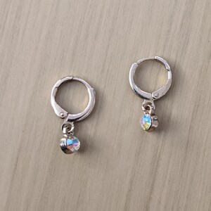 May include: A pair of silver-toned hoop earrings with a small, iridescent, round charm dangling from each. The charms reflect a rainbow of colors. The earrings are displayed on a light brown wooden surface.