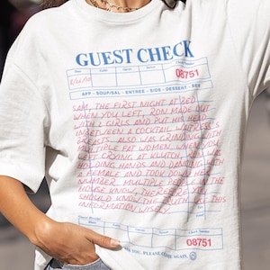 Peut inclure: T-shirt blanc avec un motif "Guest Check" bleu et rose. Le texte comprend une liste d'événements et un numéro de chèque. Le modèle porte des lunettes de soleil noires.