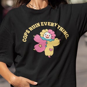 May include: Black t-shirt featuring the text "COPS RUIN EVERYTHING" in yellow, and a cartoon clown illustration. The clown is pink, yellow, and white. The t-shirt is a casual, short-sleeved style.