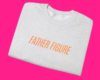 May include: A light gray sweatshirt with the words "FATHER FIGURE" printed in orange across the front. The sweatshirt is folded and set against a bright pink background. The text is in a bold, sans-serif font.