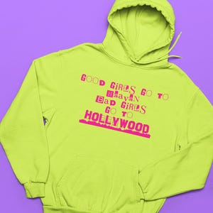 Puede incluir: Una sudadera con capucha verde neón con un bolsillo delantero y una capucha. El texto "GOOD GIRLS GO TO HEAVEN, BAD GIRLS GO TO HOLLYWOOD" está impreso en rosa. El fondo es morado.