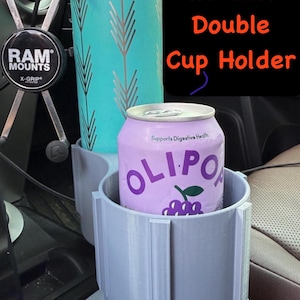 May include: A gray double cup holder for a 5th Gen 4Runner. The holder has two circular openings, one holding a teal water bottle with a silver feather design and the other holding a purple can of Olipop. The text "5th Gen 4Runner Double Cup Holder" is visible.