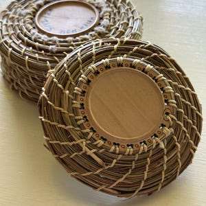 May include: Two handcrafted, round baskets made of woven natural fibers. Each basket has a wooden base and a woven rim. The baskets are light brown and beige, with a rustic, textured appearance. The baskets are likely used for storage or decoration.