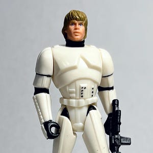 Op de afbeelding: Vintage actiefiguur van een personage uit een sciencefictionfilm. De figuur draagt een wit en zwart uniform en houdt een zwarte blaster vast. Een witte helm ligt op de grond. De figuur heeft blond haar en een strenge uitdrukking.