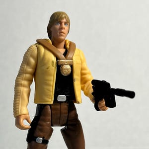 Op de afbeelding: Een plastic actiefiguur van een personage uit een sciencefictionfilm. De figuur draagt een gele jas, bruine broek, zwarte laarzen en een zwart shirt. De figuur houdt een zwart blasterpistool vast.