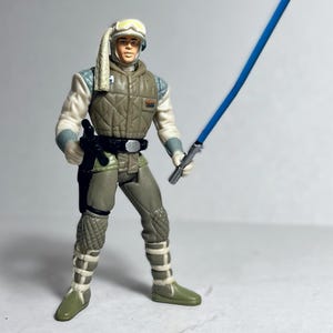 Op de afbeelding: Een Star Wars actiefiguur van een personage in een beige en witte outfit, met een blauw lichtzwaard. De figuur heeft een helm met gele bril en een zwarte riem. De figuur is ongeveer 10 cm hoog.