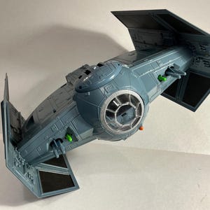 May include: A grey TIE fighter model with black solar panels. The toy spaceship has a detailed cockpit and green and orange accents. The wings are angled, and the model is set against a white background.