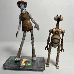 Könnte beinhalten: Zwei Actionfiguren von Außerirdischen. Eine Figur ist humanoidenartig mit brauner Weste und Hut. Die andere Figur ist ein Roboter mit rundem Torso und dünnen Gliedmaßen. Beide Figuren stehen auf einer transparenten Kunststoffbasis.