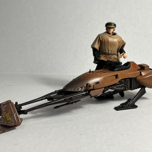 Op de afbeelding: Een bruine en zwarte speelgoed speeder bike met een figuur van een persoon in een beige outfit. De speeder bike heeft een strak ontwerp met lange, dunne verlengingen aan de voorkant en een gedetailleerde motor. De figuur staat naast de speeder bike.