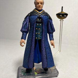 Könnte beinhalten: Detaillierte Actionfigur einer Figur in einem langen blauen und lila Gewand mit goldenen Akzenten. Die Figur steht auf einer transparenten Basis und hat ein kleines, schwarz-goldenes Gerät auf einem transparenten Ständer. Graues Haar.