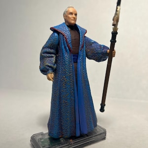 Könnte beinhalten: Detaillierte Actionfigur einer Figur in einem langen blauen Gewand mit goldenen Akzenten, die einen Stab hält. Die Figur steht auf einer transparenten Kunststoffbasis. Das Gewand hat ein strukturiertes Muster und rote Verzierungen.