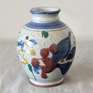 May include: A small, round ceramic vase with a speckled off-white glaze. The vase features hand-painted floral motifs in shades of blue, red, yellow, and green. It has a narrow neck with a blue rim and a flared base, showcasing a rustic, textured finish.