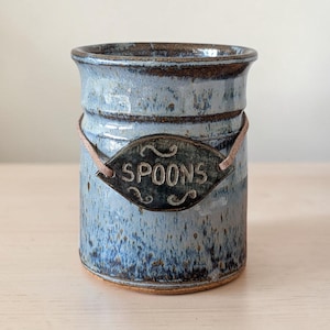 May include: A cylindrical ceramic utensil holder with a speckled blue and brown glaze. A dark, diamond-shaped clay plaque is attached to the front with a thin leather cord, featuring the embossed text SPOONS and decorative scrollwork details.