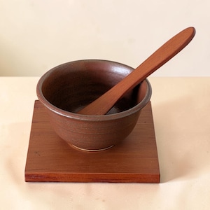 May include: A dark brown ceramic bowl with a smooth, rounded rim sits centered on a square wooden base. A long, flat wooden spoon with a tapered handle rests inside the bowl, extending diagonally upward from the interior base to the outer edge.