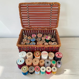May include: A rectangular woven basket with a hinged lid containing a collection of assorted sewing thread spools. Additional spools of various colours, sizes, and brands, including Coats and Singer, are arranged in front of the basket on a surface.