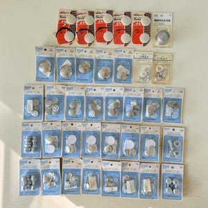 May include: A collection of various sewing button kits on cardboard backing cards, including Scovill and Unique brands. The packages contain metal button components, plastic backings, and assembly tools, arranged in rows on a white surface.