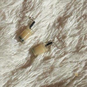 Op de afbeelding: Een paar novelty oorbellen in de vorm van miniatuur bubble tea. Elke oorbel heeft een doorzichtige, rechthoekige beker gevuld met een beige vloeistof, zwarte boba en een zwart rietje. De oorbellen hebben zilverkleurige oorhaaksluitingen.