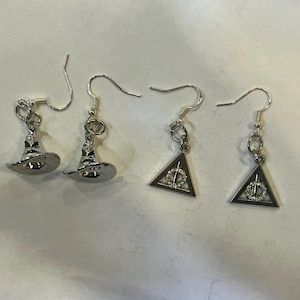 May include: Silver-toned earrings featuring two pairs of charms. One pair showcases miniature witch hats, while the other displays a triangle with a circular design. The earrings have a hook closure.